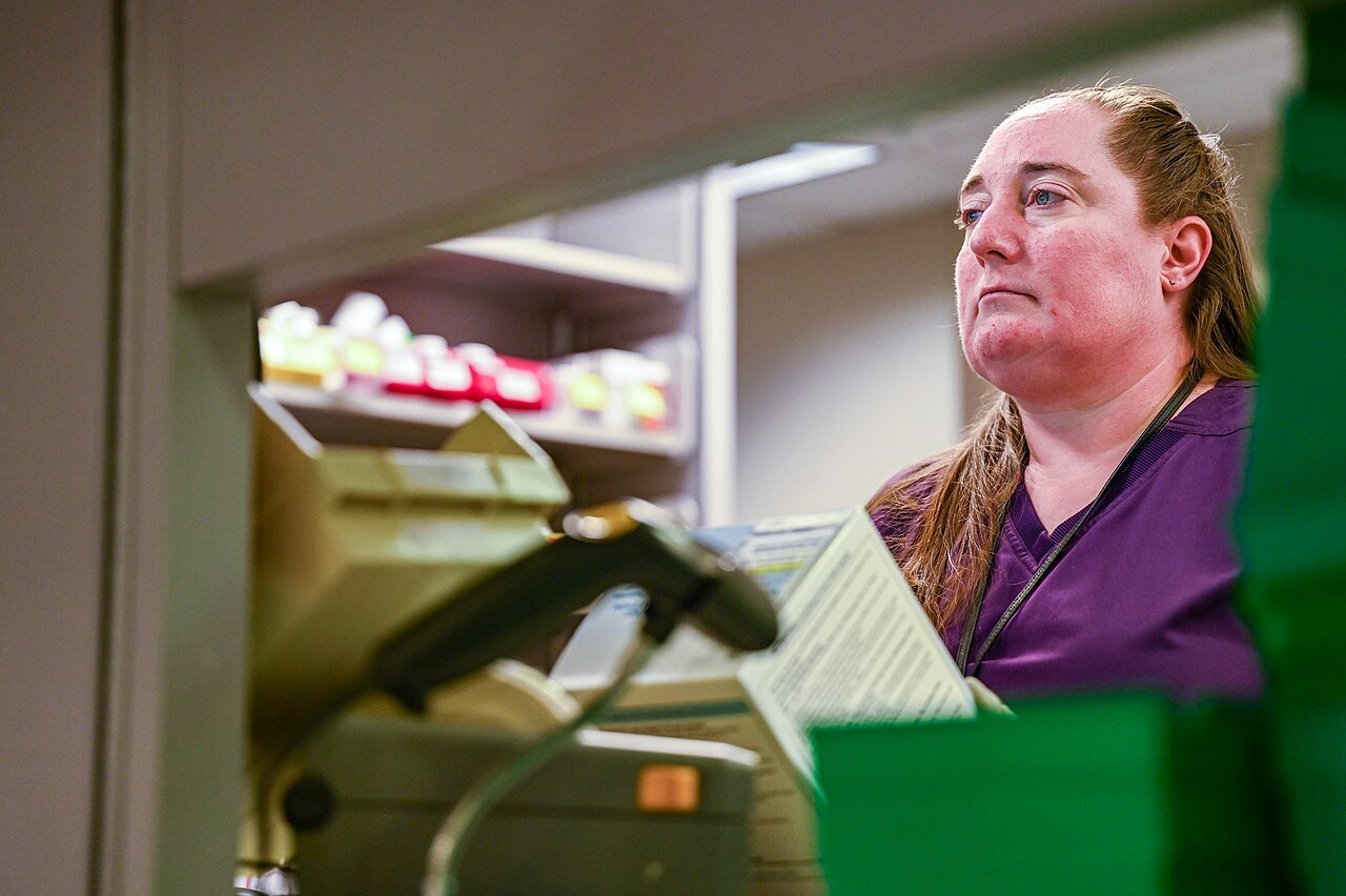 A pharmacy technician in purple scrubs scans medication at a pharmacy counter.